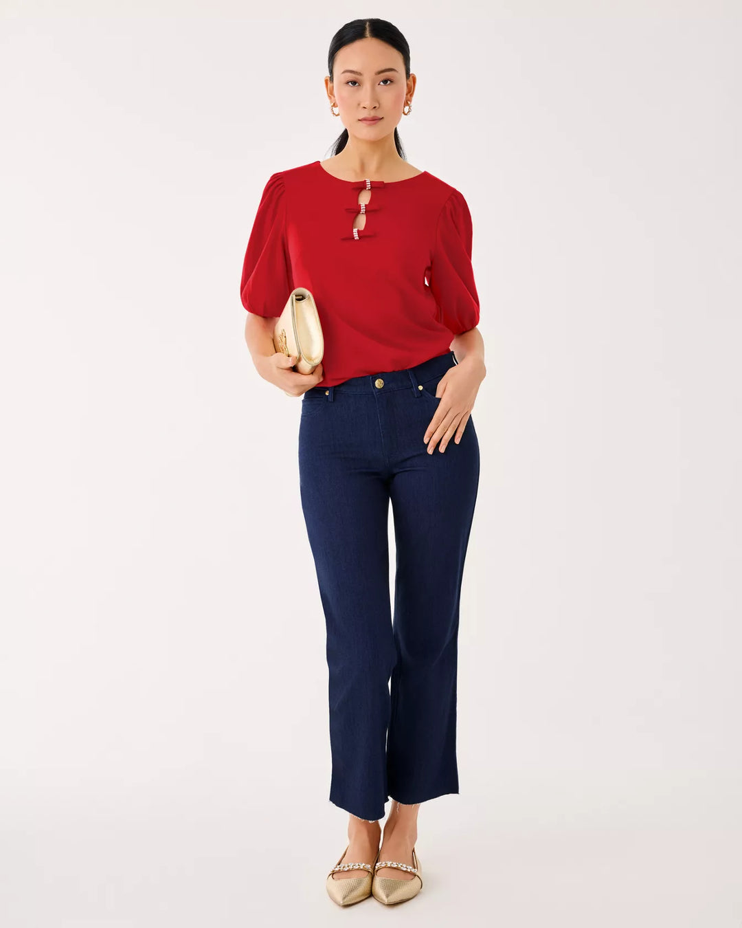Woman wearing a red blouse and navy pants holding a beige clutch against a white background