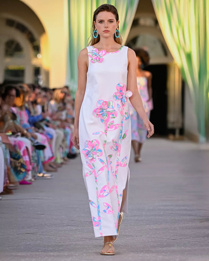 Model walking on a fashion show runway wearing a white dress with colorful floral patterns.