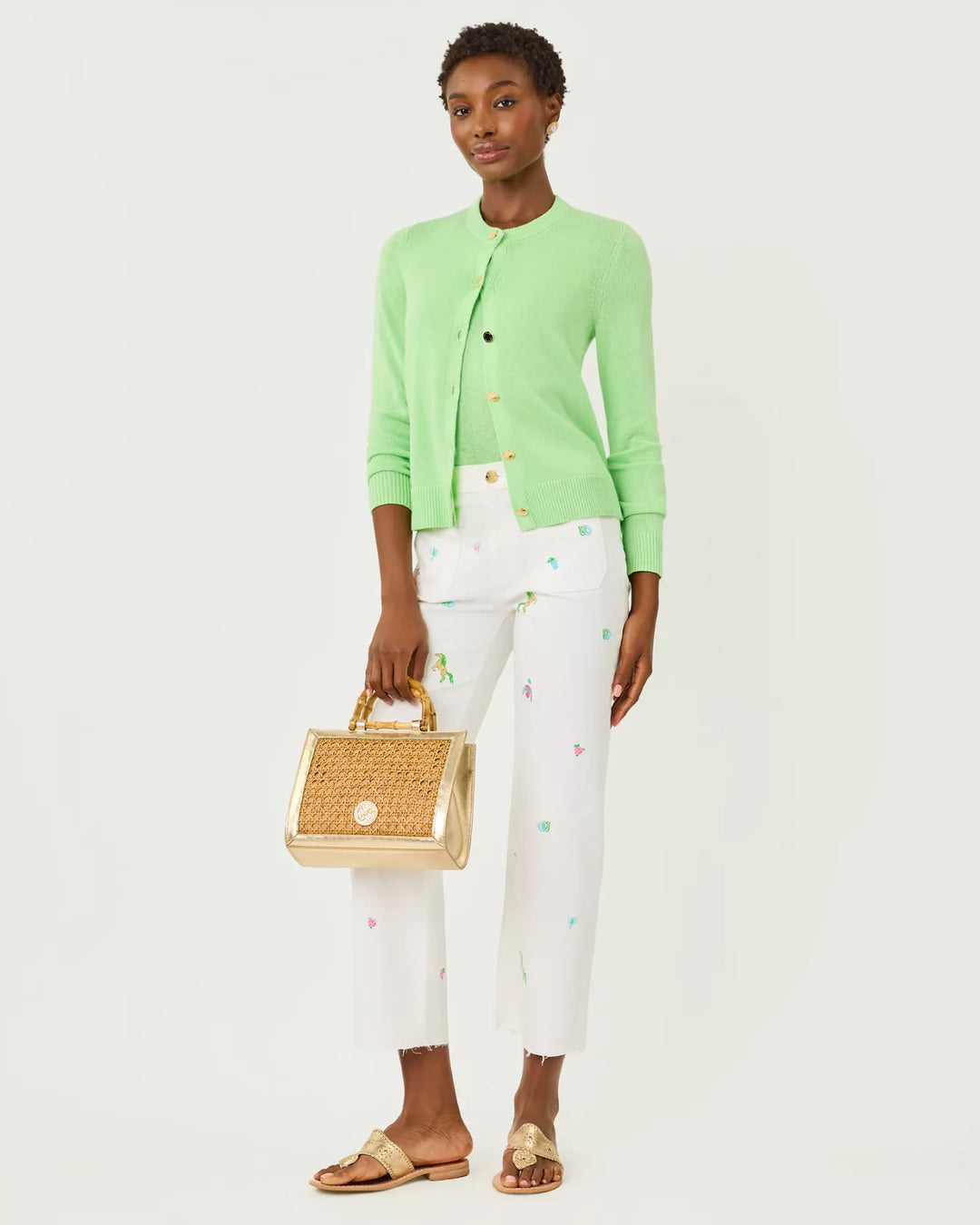 Woman wearing a green cardigan and white pants holding a gold handbag on a white background