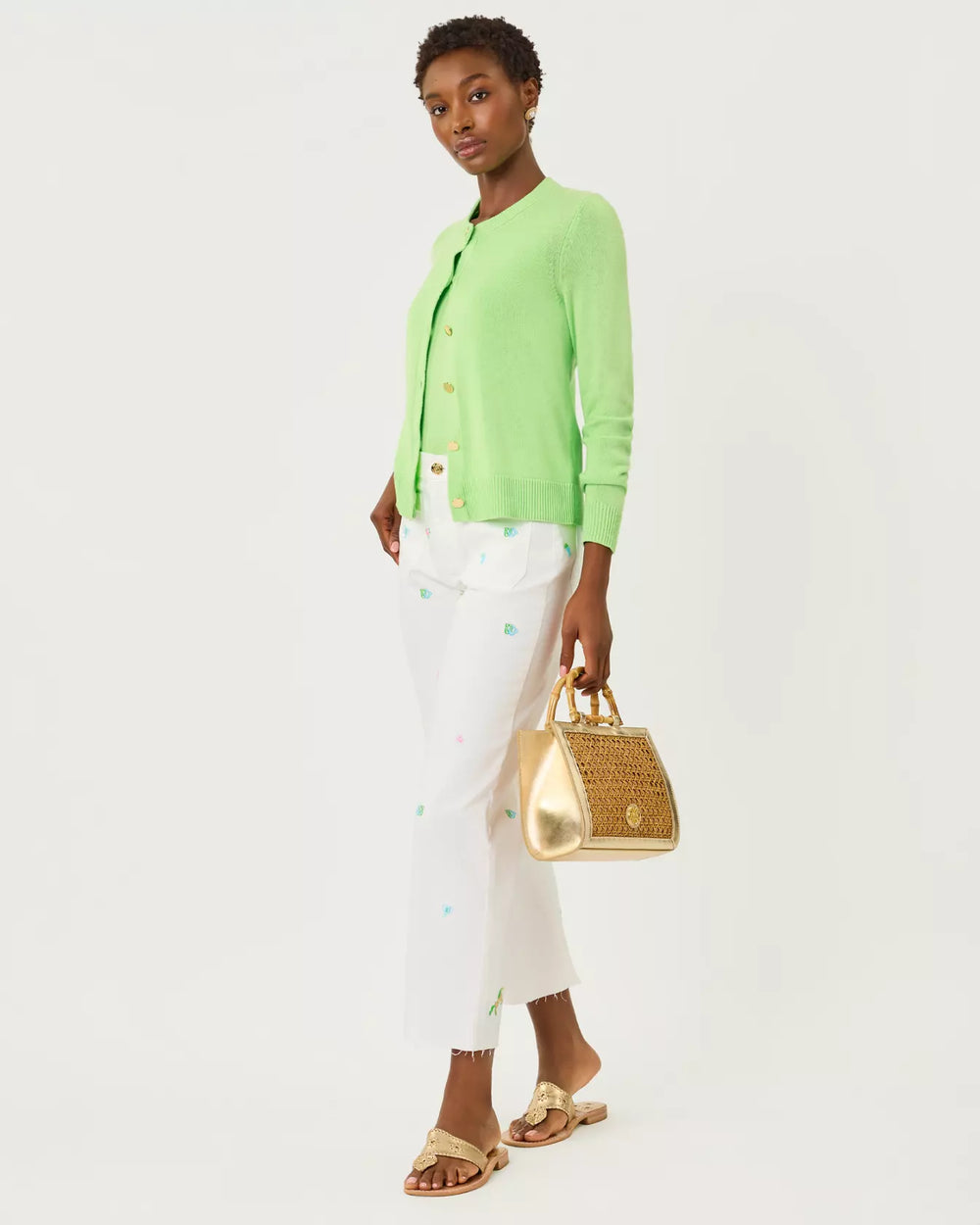 Woman wearing a light green cardigan, white pants, and gold sandals holding a gold handbag on a white background